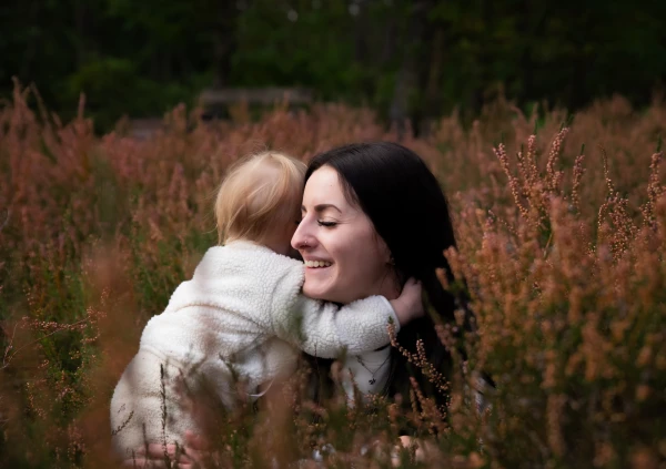 Mutter liegt in der Lüneburger Heide auf dem Bauch und wird von ihrer Tochter umarmt. Man sieht das Kind nur von hinten.